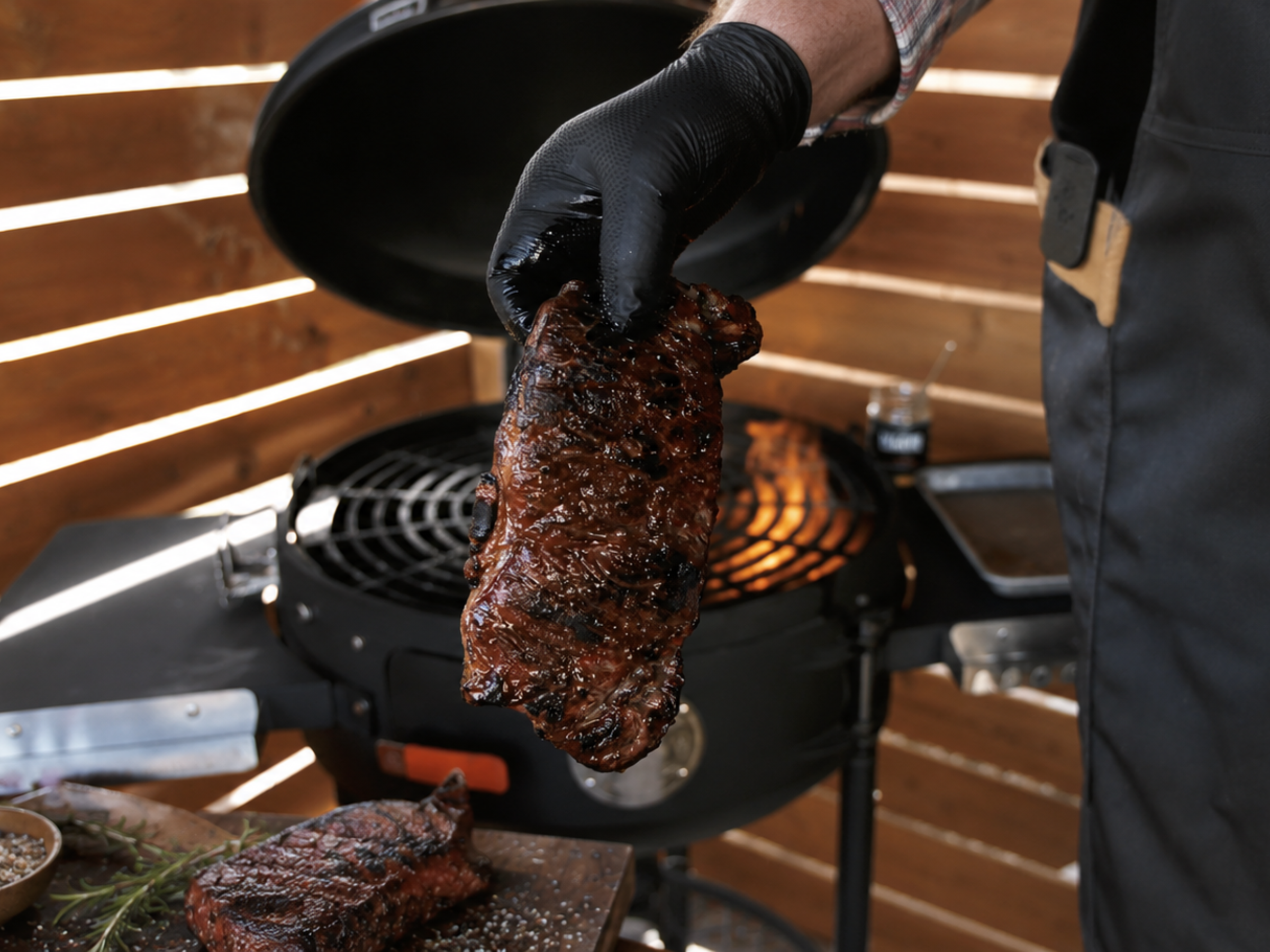 A person wearing a black glove holds up a well-charred ribeye steak in front of an open black grill with visible flames. Another steak sits on a cutting board in the foreground, with a wooden slatted wall in the background.